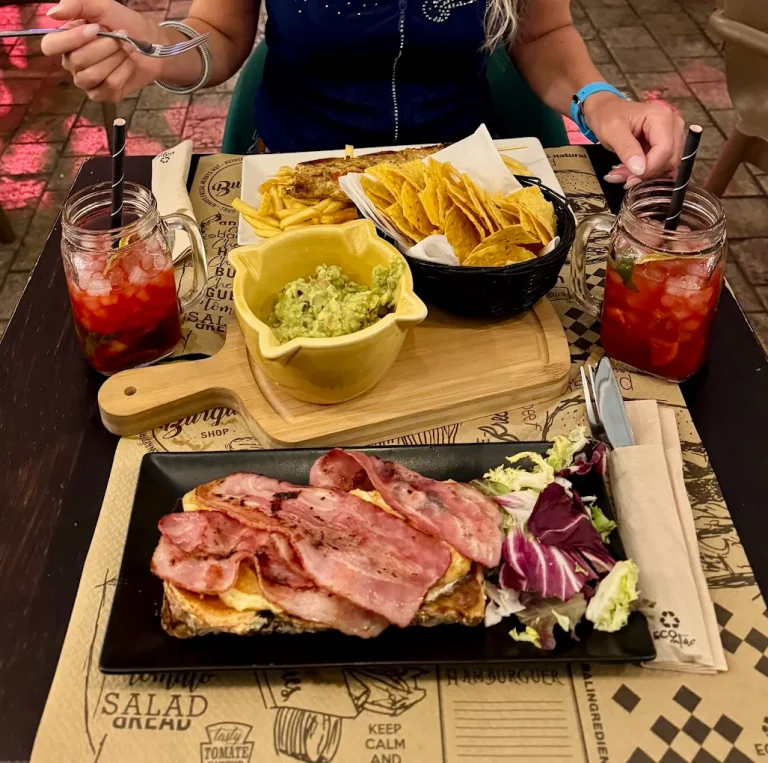 Plato con sándwich de jamón y ensalada, acompañados de nachos y guacamole. Bebidas rojas al fondo.