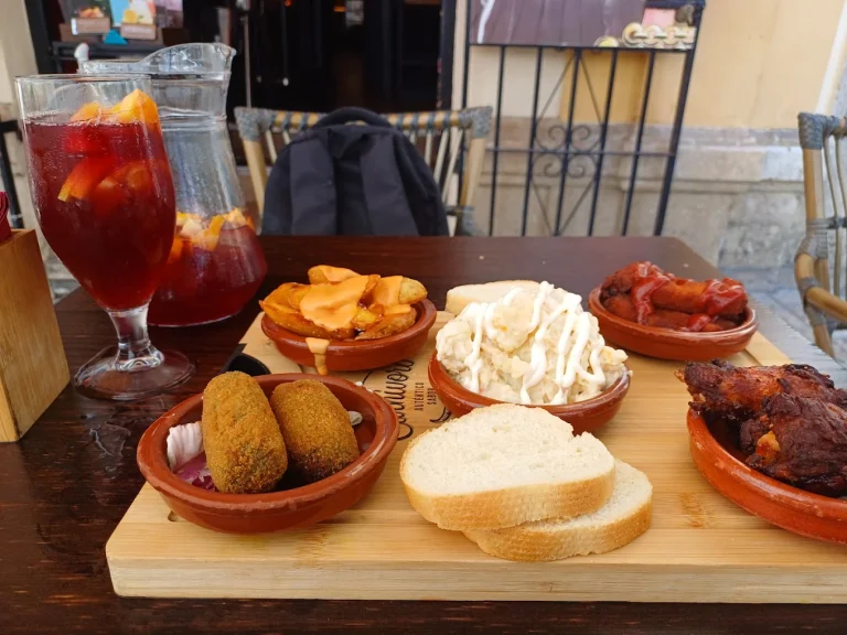 Tapa variada con croquetas, alitas, patatas y pan, acompañada de sangría en dos vasos.