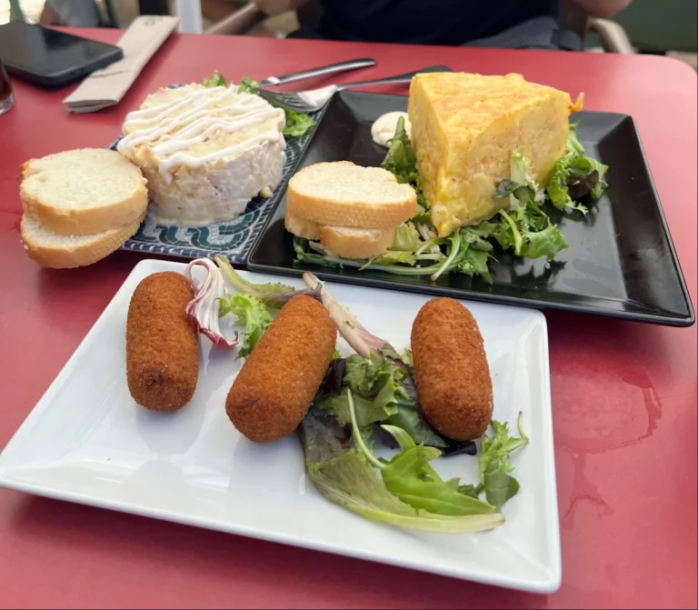 Platos con croquetas, ensalada, pan y un pastel amarillo en la mesa.